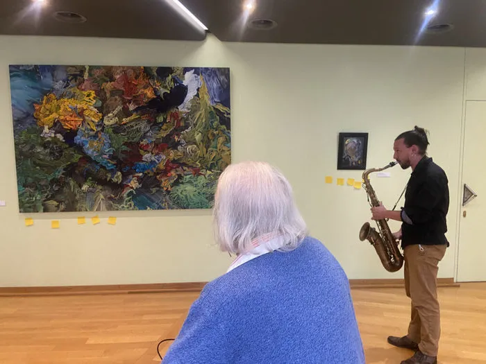 Une personne âgée regarde un tableau coloré au mur, un saxophoniste joue de son instrument.