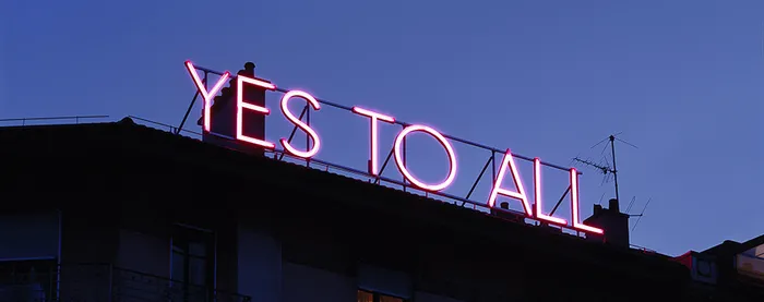 Néon rose avec inscription "Yes to all" au somment d'un immeuble à la tombée de la nuit.